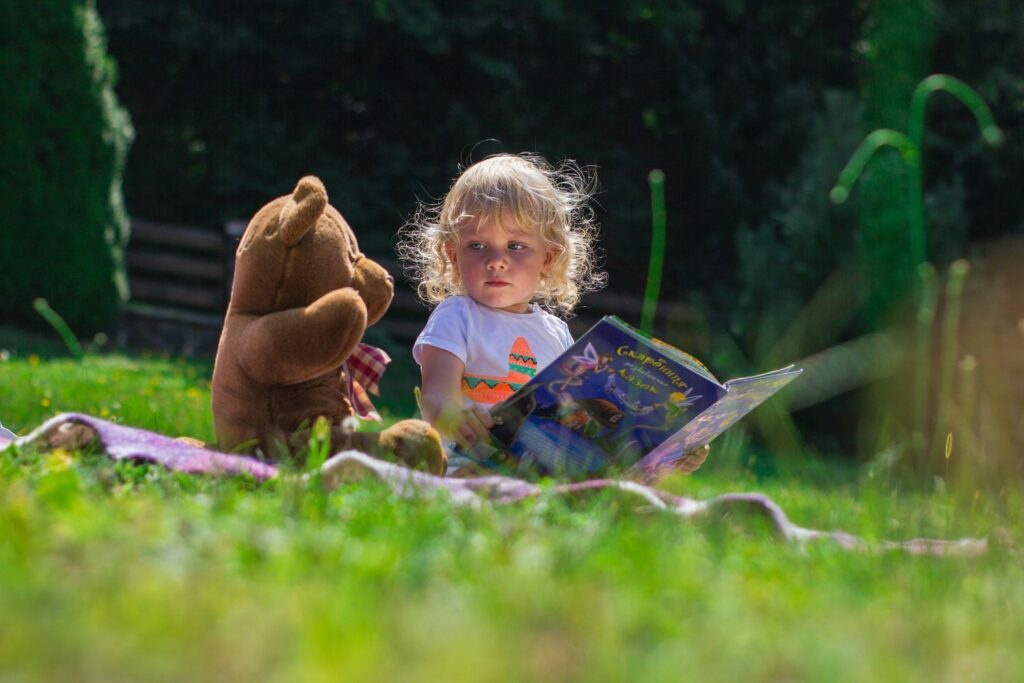 kleines Mädchen mit großem Plüschbär auf der Wiese liest aus Märchenbuch vor