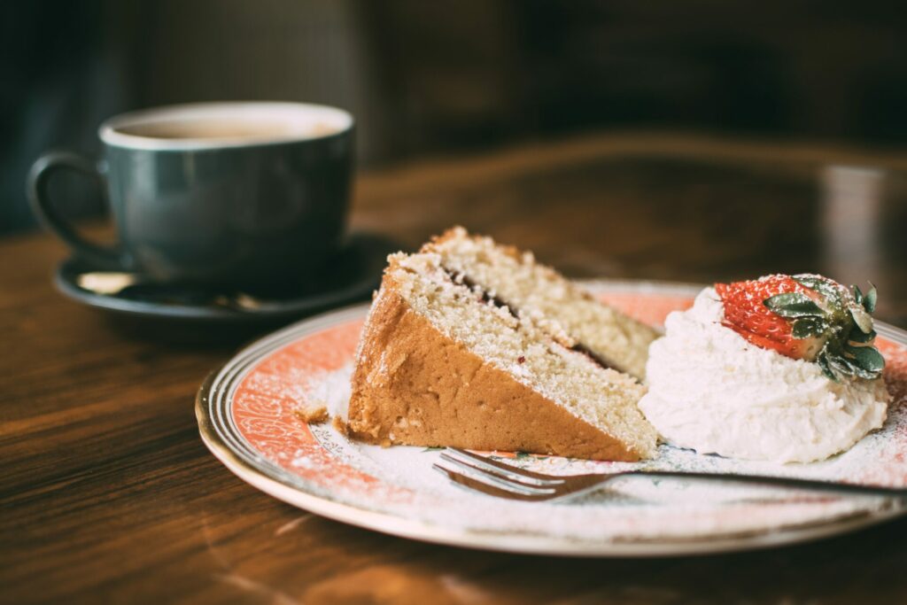 leckeres Stück Kuchen auf einem Teller mit Kuchengabel einen Klecks Sahne auf dem eine Erdbeere liegt und eine Tasse kaffee
