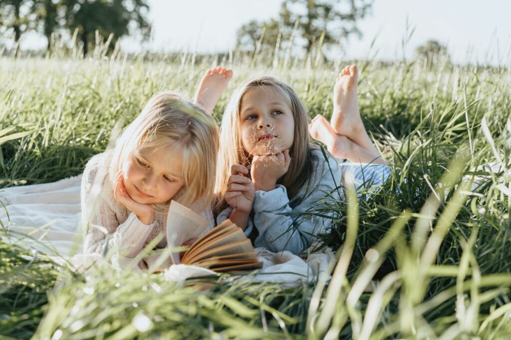 zwei Mädchen liegen auf einer Wiese, eine träumt, die andere liest in einem buch