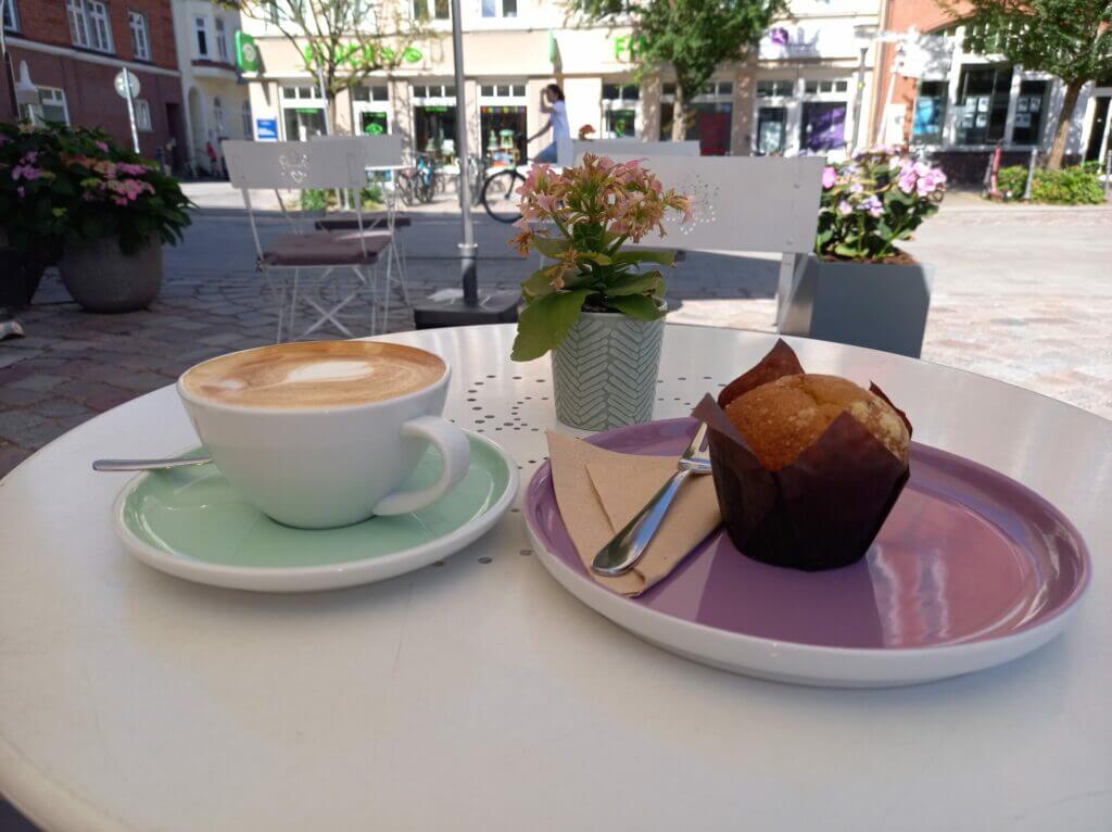 Weißes Metaltischchen mit einer Tasse Cappuccino und einem Kuchenteller mit einem Muffin drauf